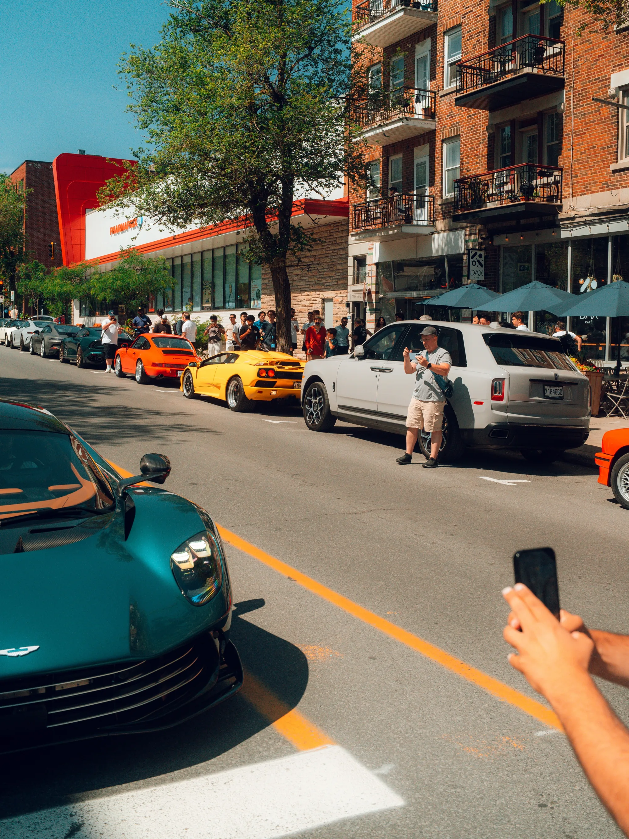 The crowd along the curb, beside a teal Aston Martin, a Rolls-Royce and a yellow Diablo, with café umbrellas up behind.