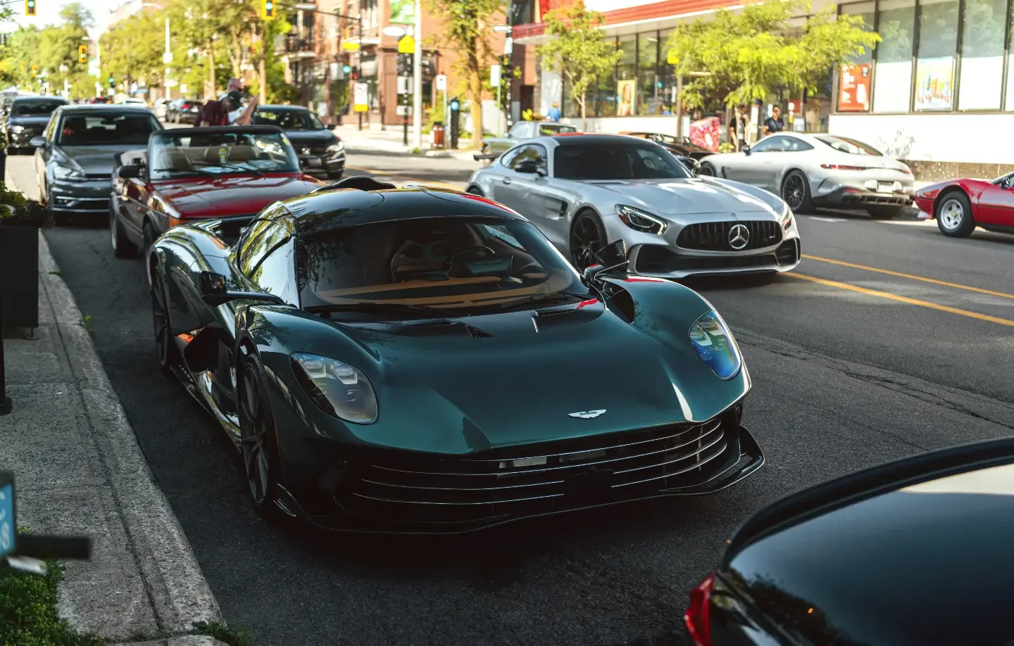 An Aston Martin Valhalla in racing green pulls up beside a silver Mercedes-AMG GT R and a red Ferrari on a Montréal street.