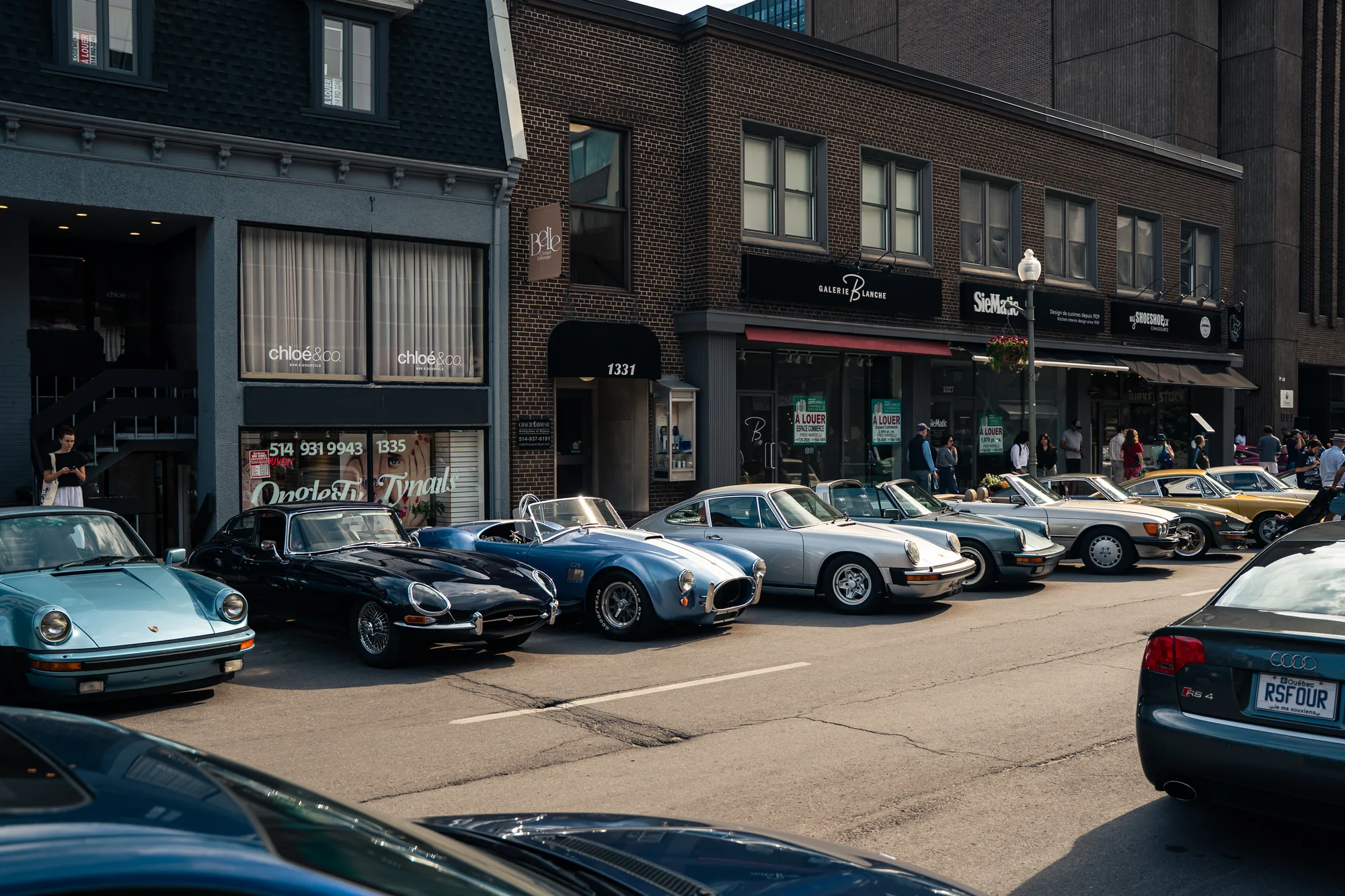 A row of classics angled at the curb on Greene Avenue: a blue Porsche 911, a dark Jaguar E-Type, a blue Cobra and two more 911s in front of the shopfronts.