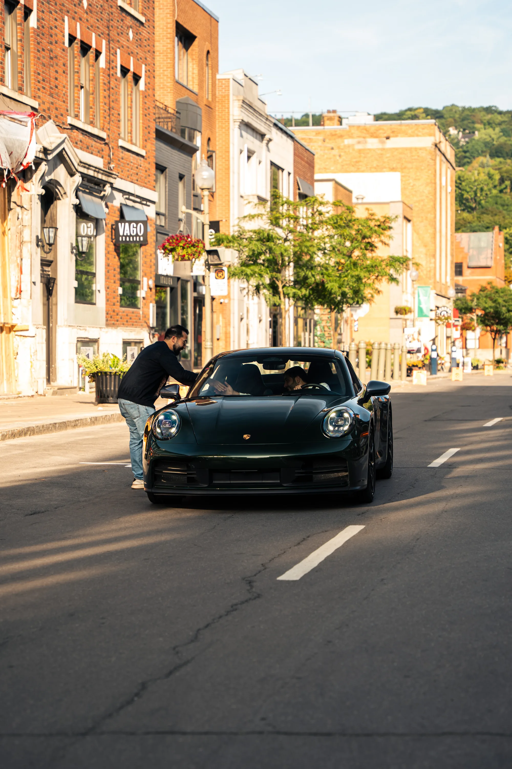 A dark green Porsche 911 pulls up Greene Avenue while a man leans in at the driver’s window.
