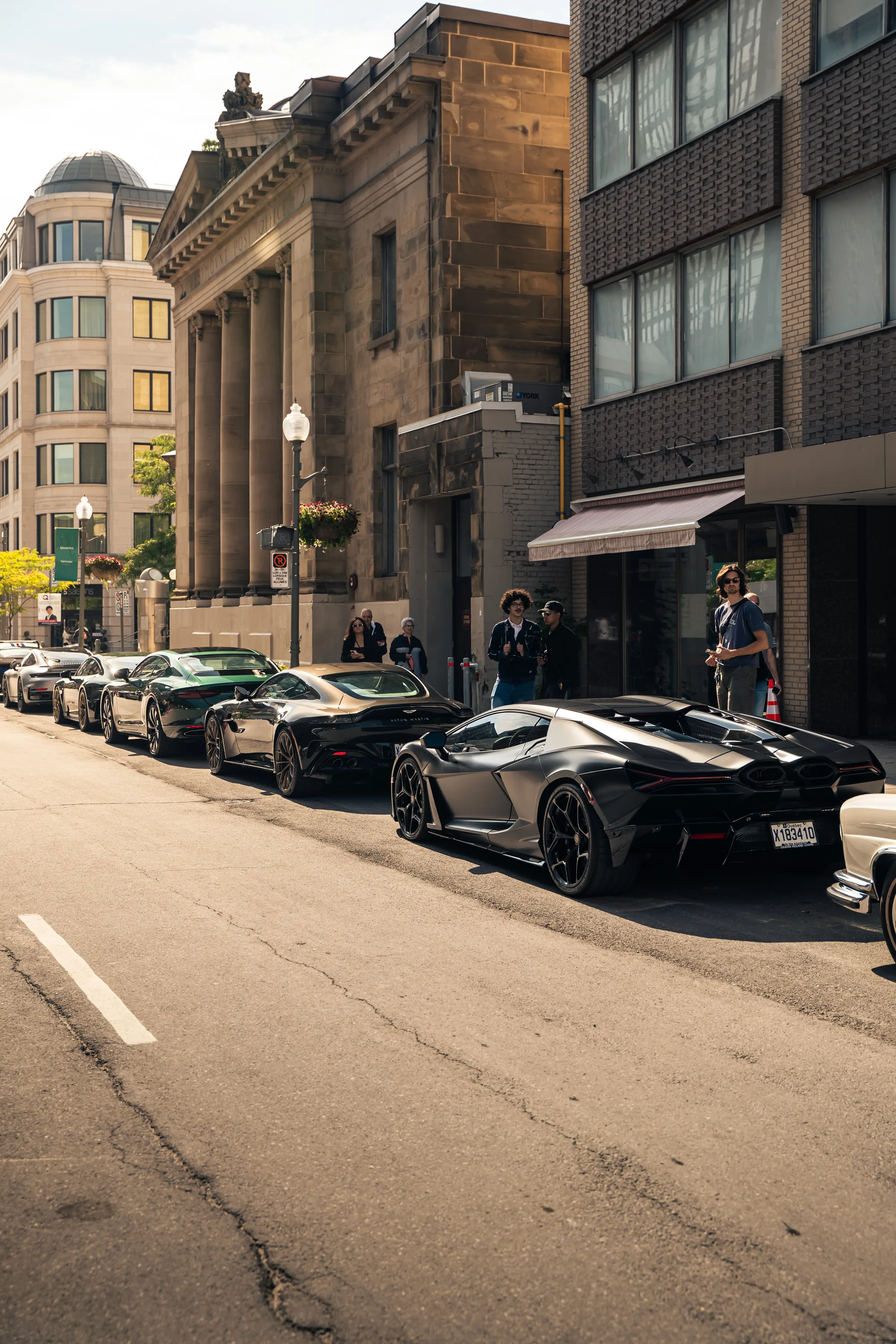 A matte black Lamborghini parked along Greene Avenue below the columned limestone bank, a black Aston Martin behind it.